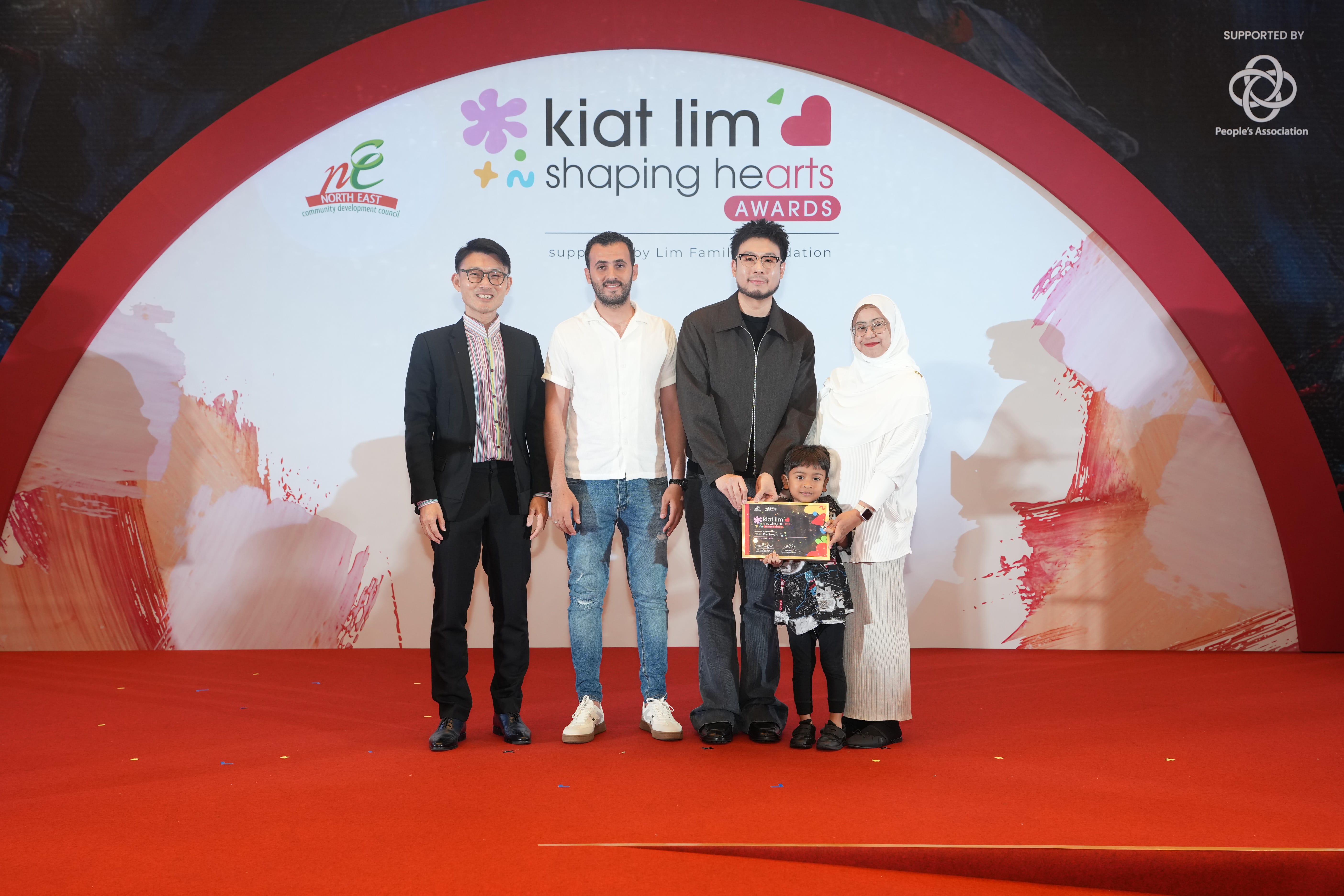 Award recipient standing on stage with the presenter, holding their certificate and posing for a group photo during the award ceremony, with a backdrop reading ‘Kiat Lim-Shaping Hearts Awards'.